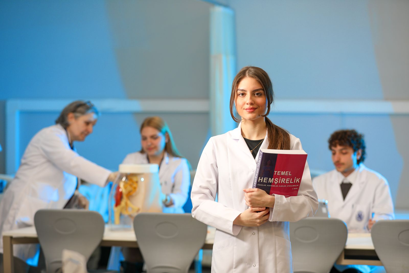 Free Young nursing students engaging in lab study, showcasing medical education. Stock Photo.Free Young woman in a lab coat holding a nursing textbook in a classroom setting.